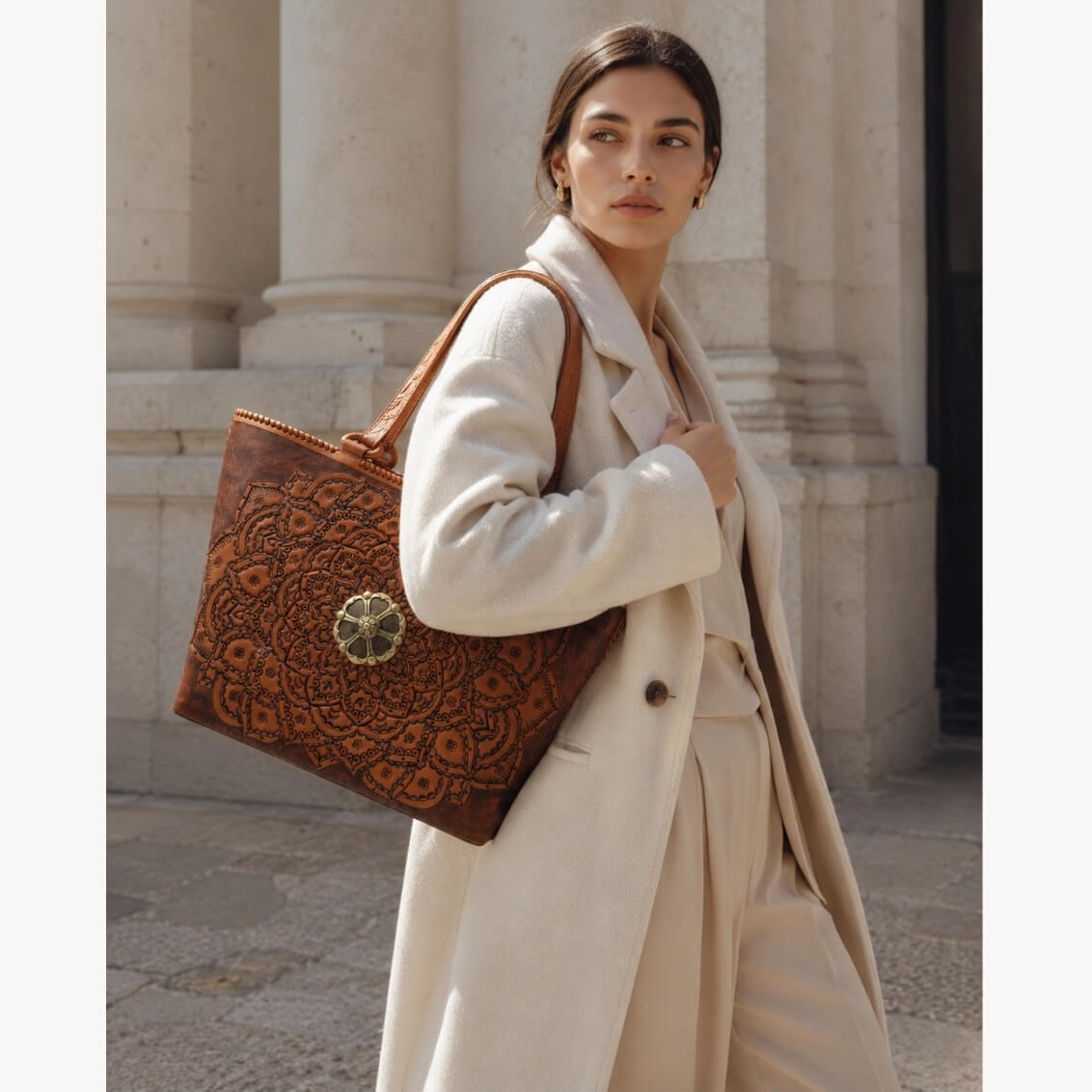 Woman in a beige coat holding a brown leather bag with intricate patterns, standing in front of classical architecture.