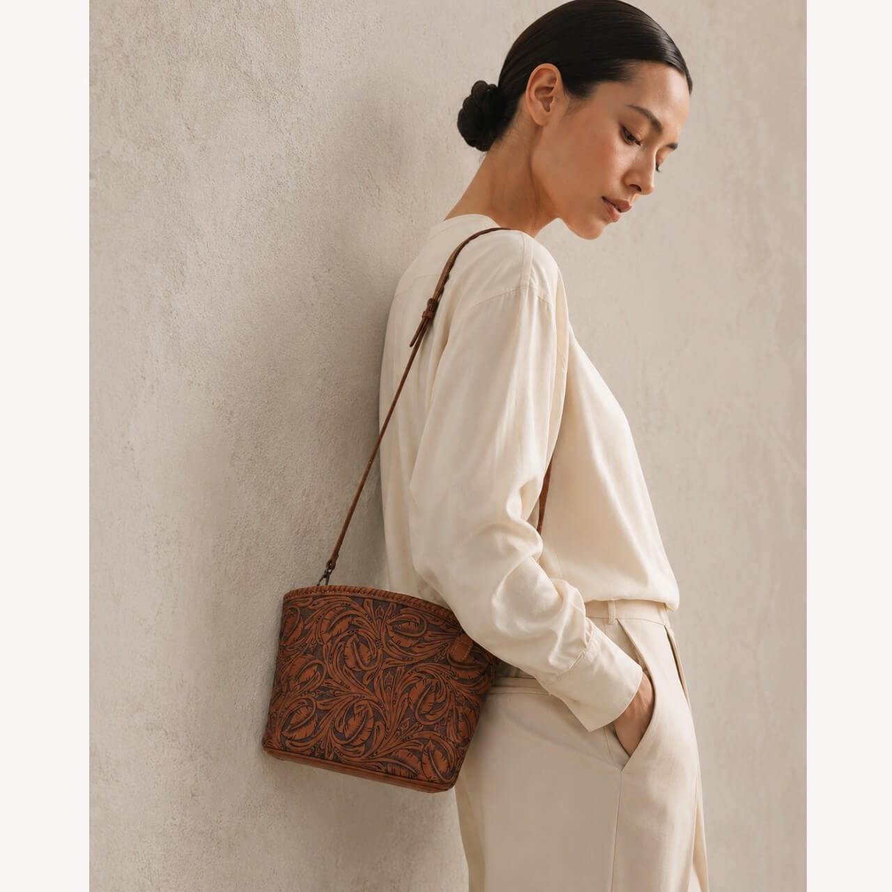 Model wearing a burnt orange hand-tooled leather shoulder bag with floral detailing and a slim leather strap.