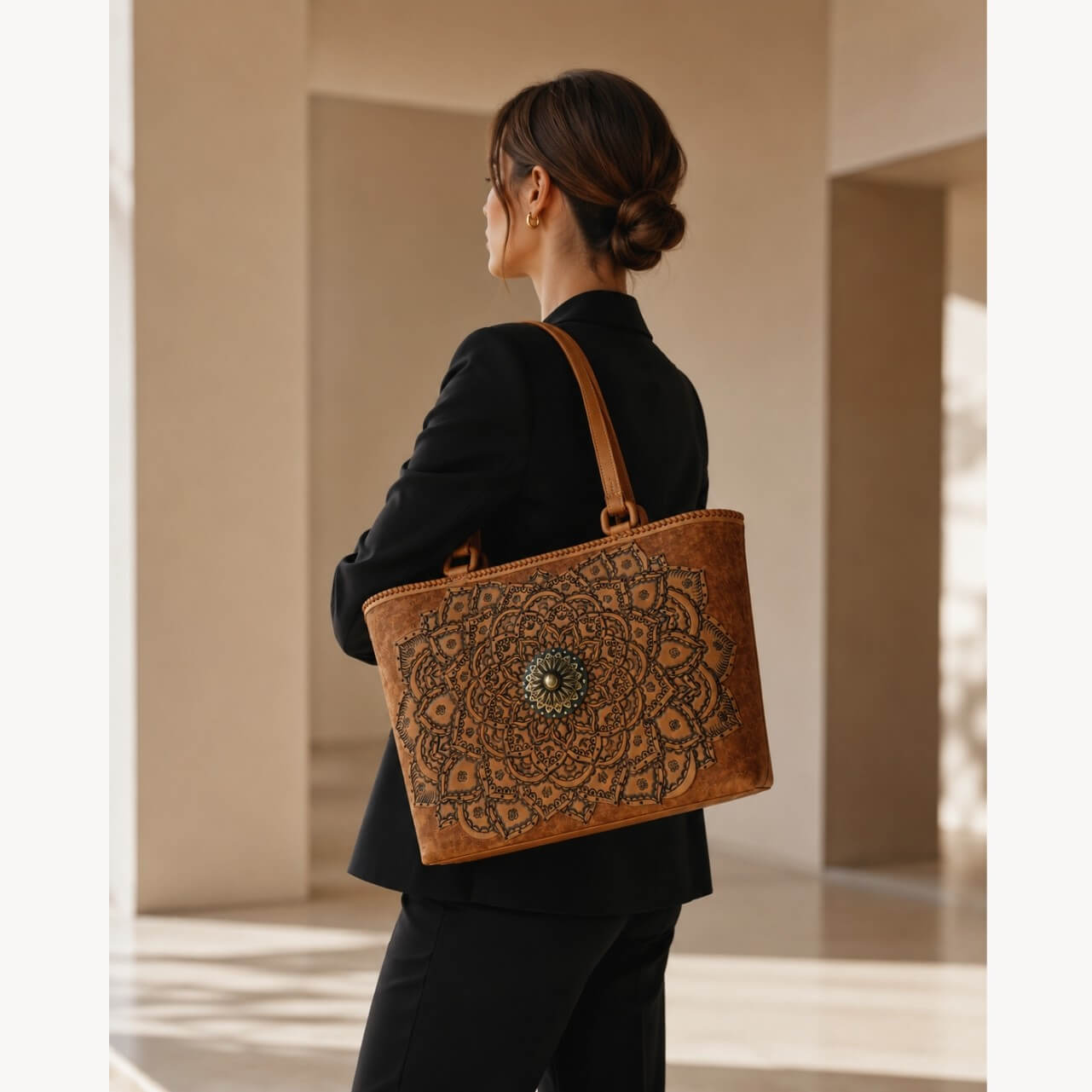Cognac/burnished brown nude color tooled leather tote with mandala tooling and antique medallion hardware carried over the shoulder by a model in a black blazer.