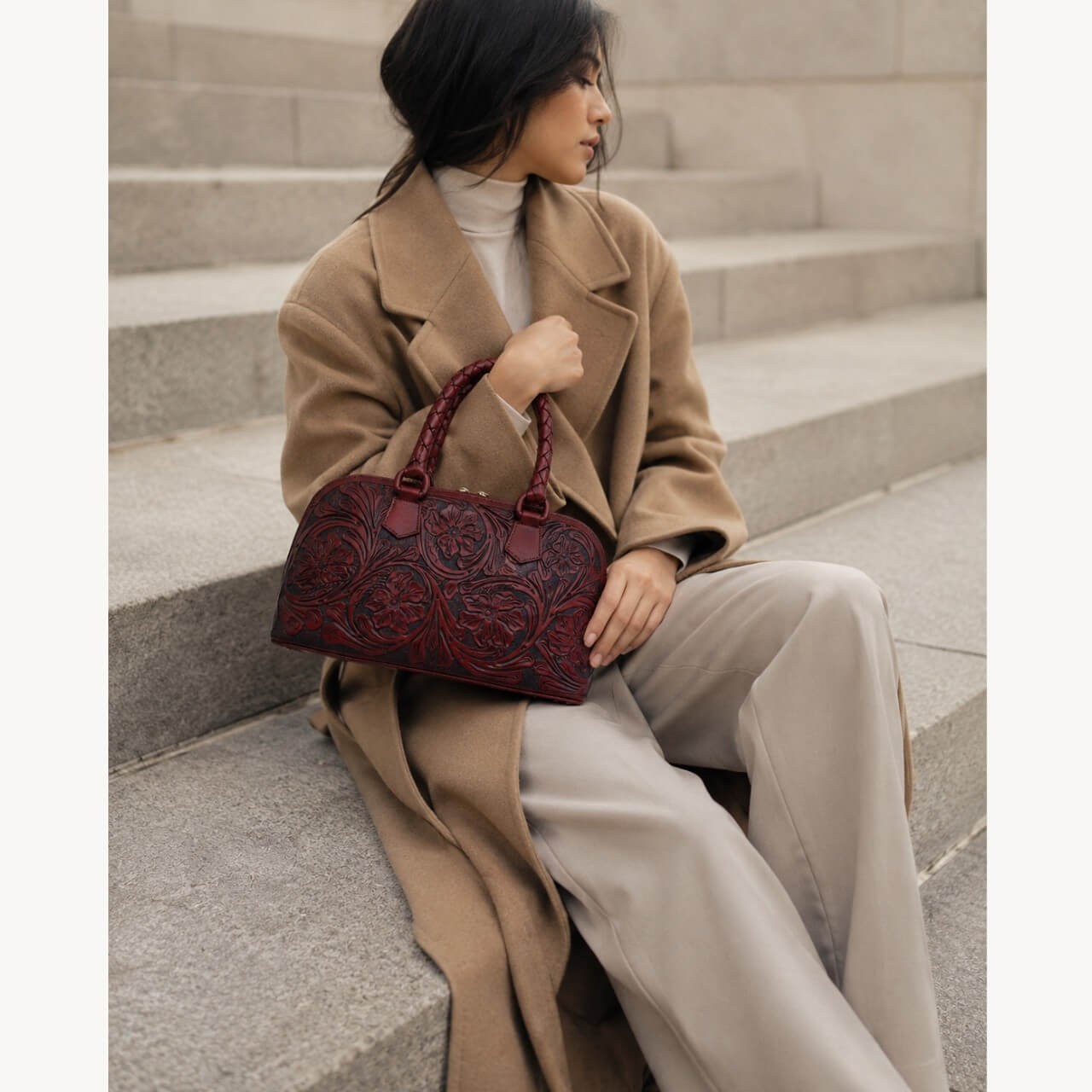 Seated model holding a Mahogany Espresso hand-tooled leather satchel bag with floral detailing and braided top handles.