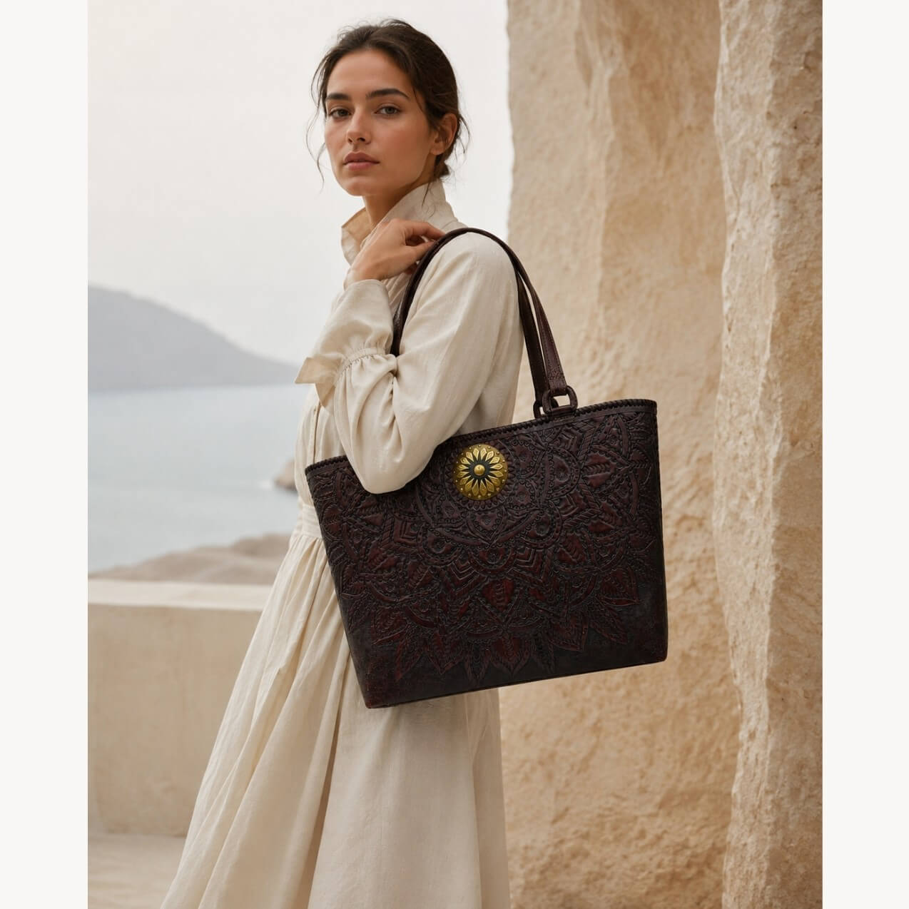 Model in a cream dress carrying a mahogany espresso mandala embossed leather tote with top handles and a round gold medallion.