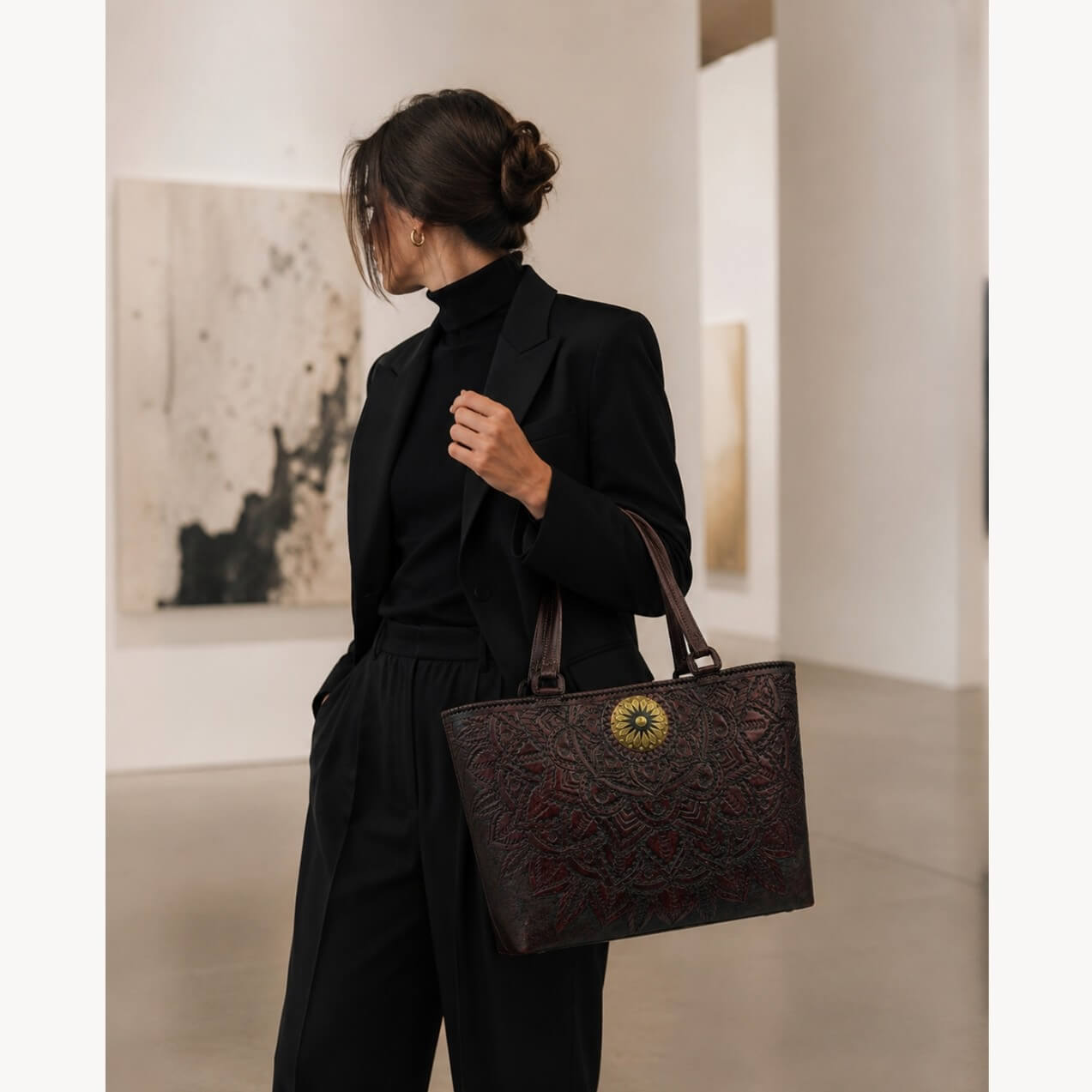 Model in a black outfit carrying a mahogany espresso mandala embossed leather tote with top handles and a round gold medallion in an art gallery.