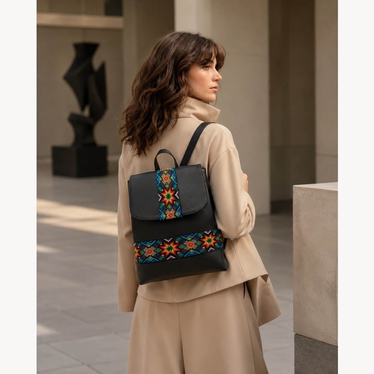 Model wearing a black leather backpack with colorful woven geometric textile panels, styled with a beige outfit in a modern architectural setting.