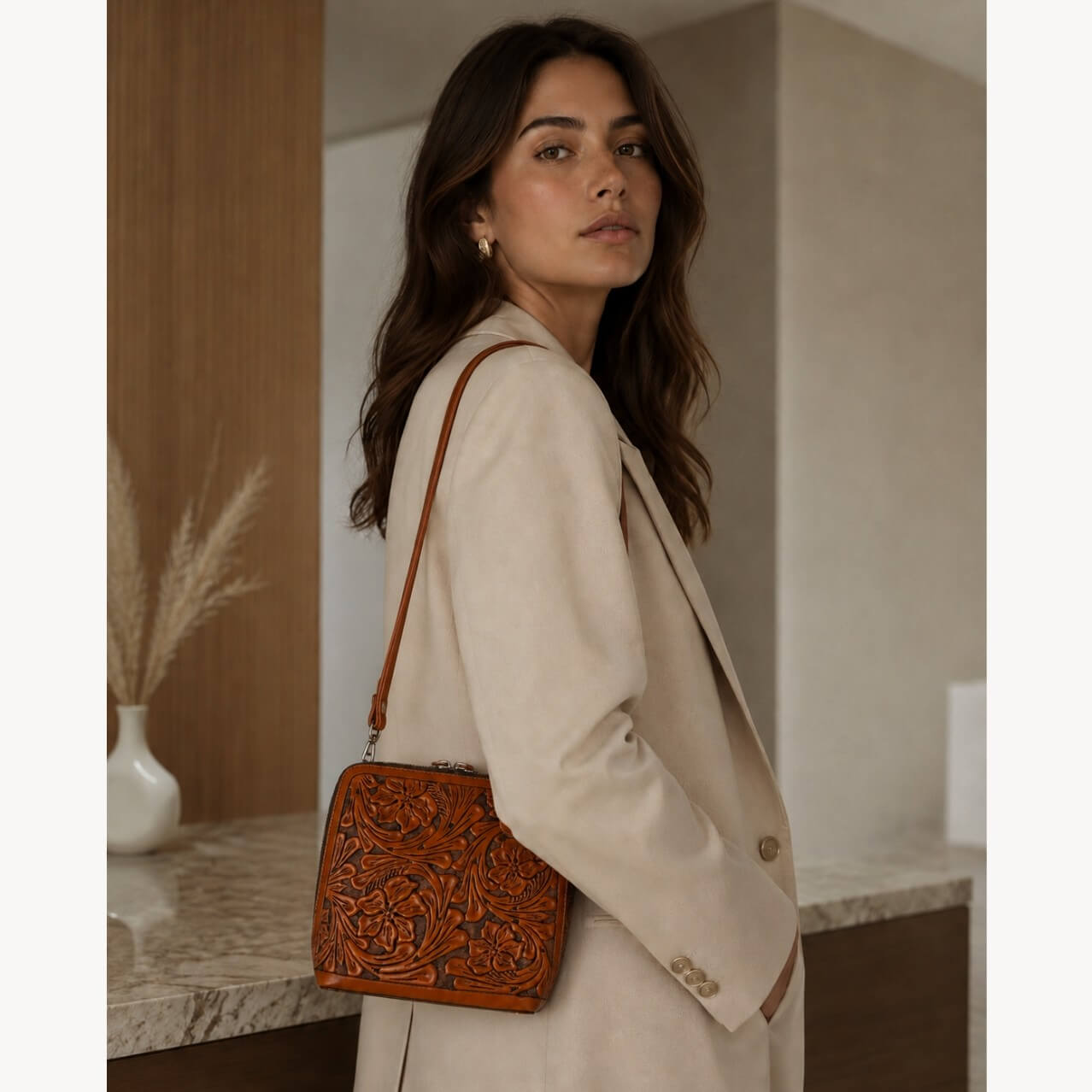 Model wearing a burnt orange hand-tooled floral leather crossbody bag with a long strap, styled with a cream blazer in a modern interior setting.