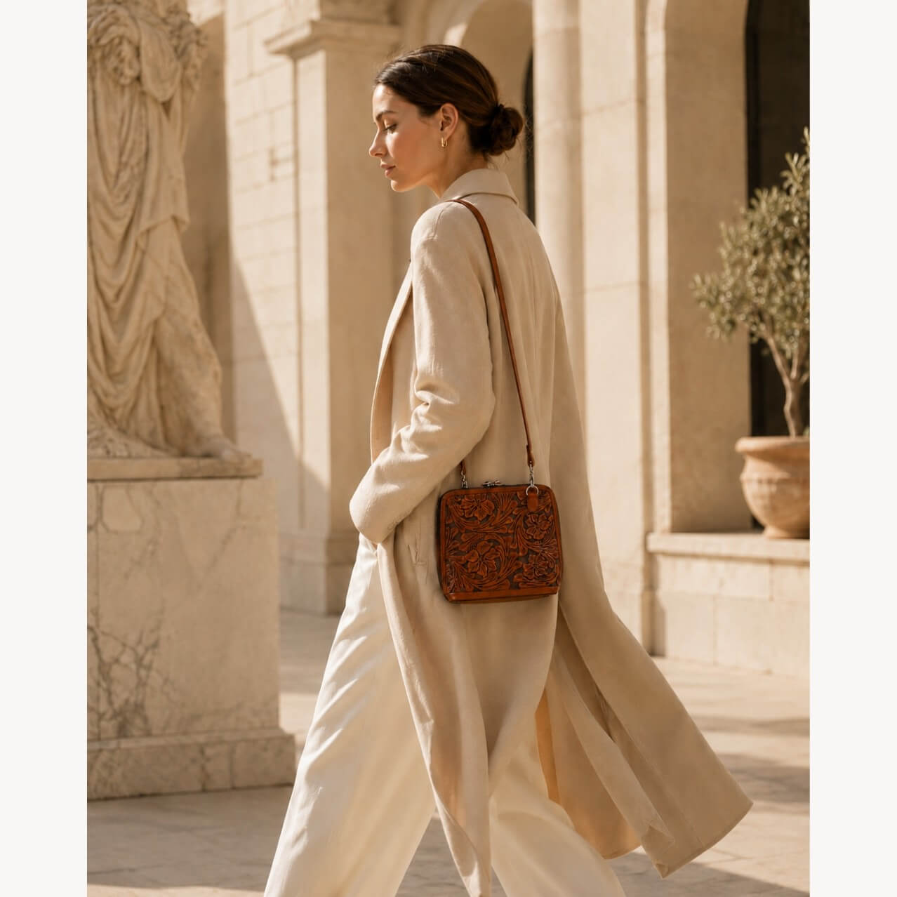 Model wearing a burnt orange hand-tooled floral leather crossbody bag with a long strap, styled with a cream coat in an architectural courtyard setting.
