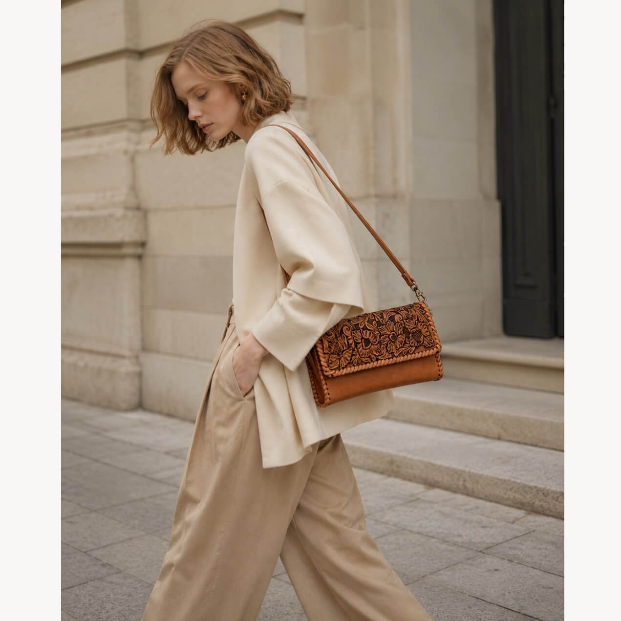 Model wearing a natural tan hand-tooled leather crossbody bag with floral detailing and a slim leather shoulder strap.