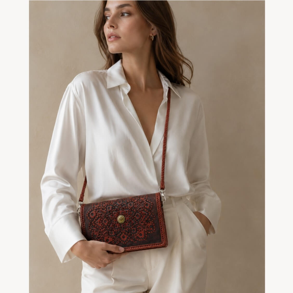 Model in a white outfit wearing a red-brown embossed leather crossbody bag with mandala detailing and a gold circular clasp.