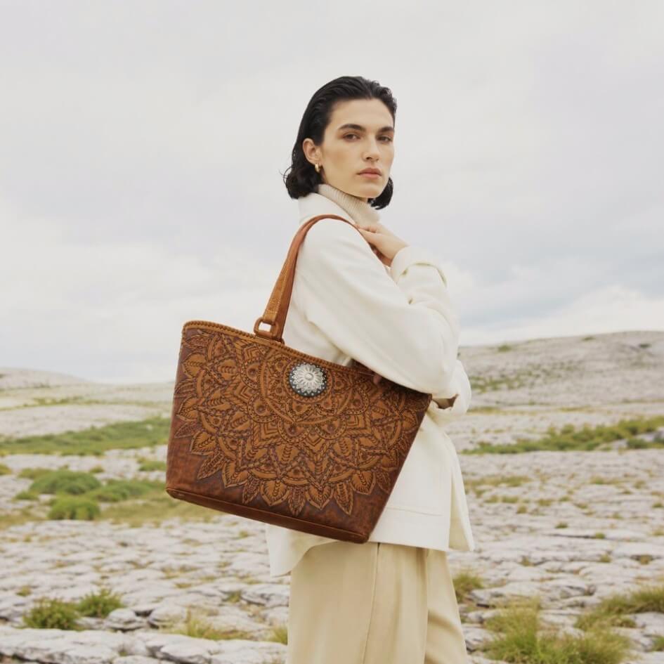 Woman holding a brown leather bag with intricate patterns in an open field.