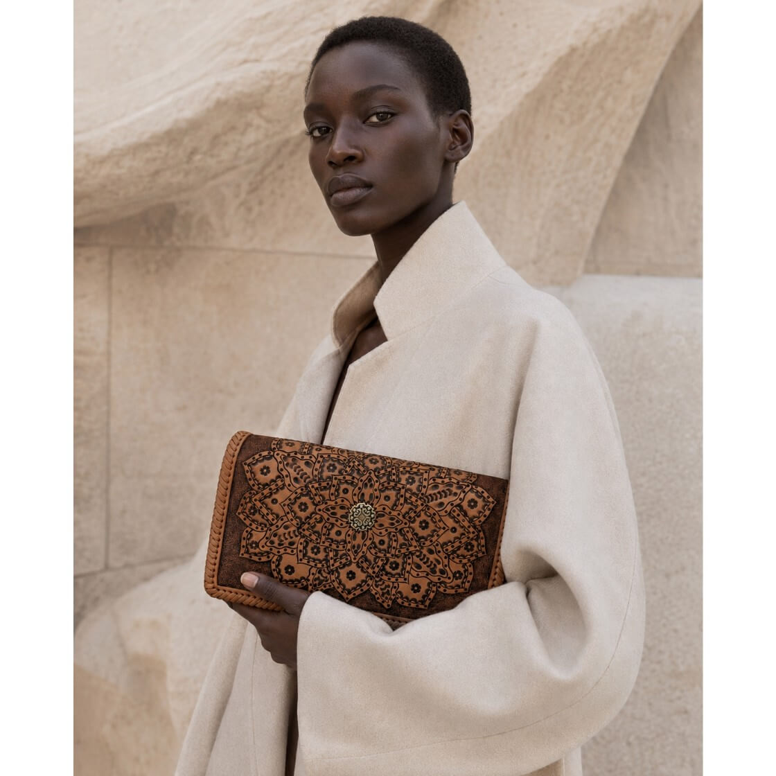 Woman holding a full grain leather clutch with floral pattern against a beige stone wall.