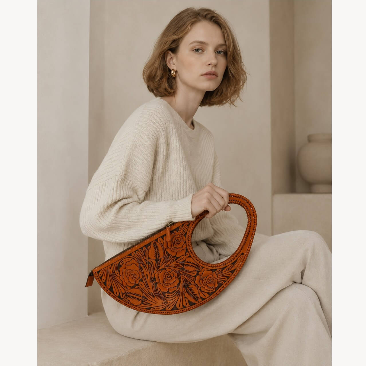 Seated model holding a burnt orange hand-tooled leather handbag with floral rose detailing and braided circular top handle.