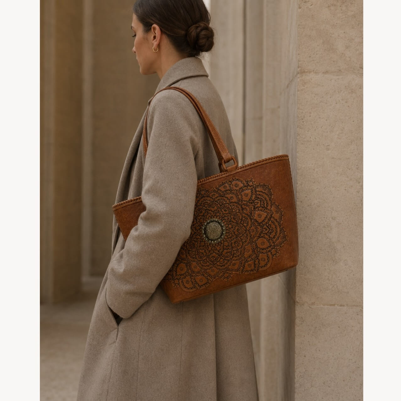 Cognac/burnished brown nude color tooled leather tote with floral medallion detail carried over the shoulder by a model in a gray coat near a stone wall.