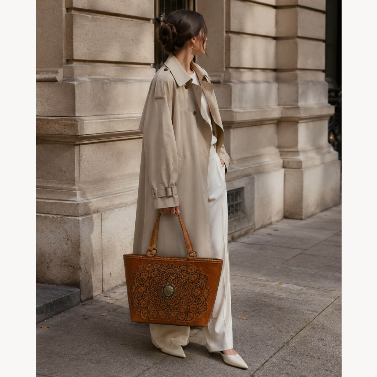 Cognac/burnished brown nude color tooled leather tote with floral medallion detail carried by a model in a beige trench coat on a city sidewalk.