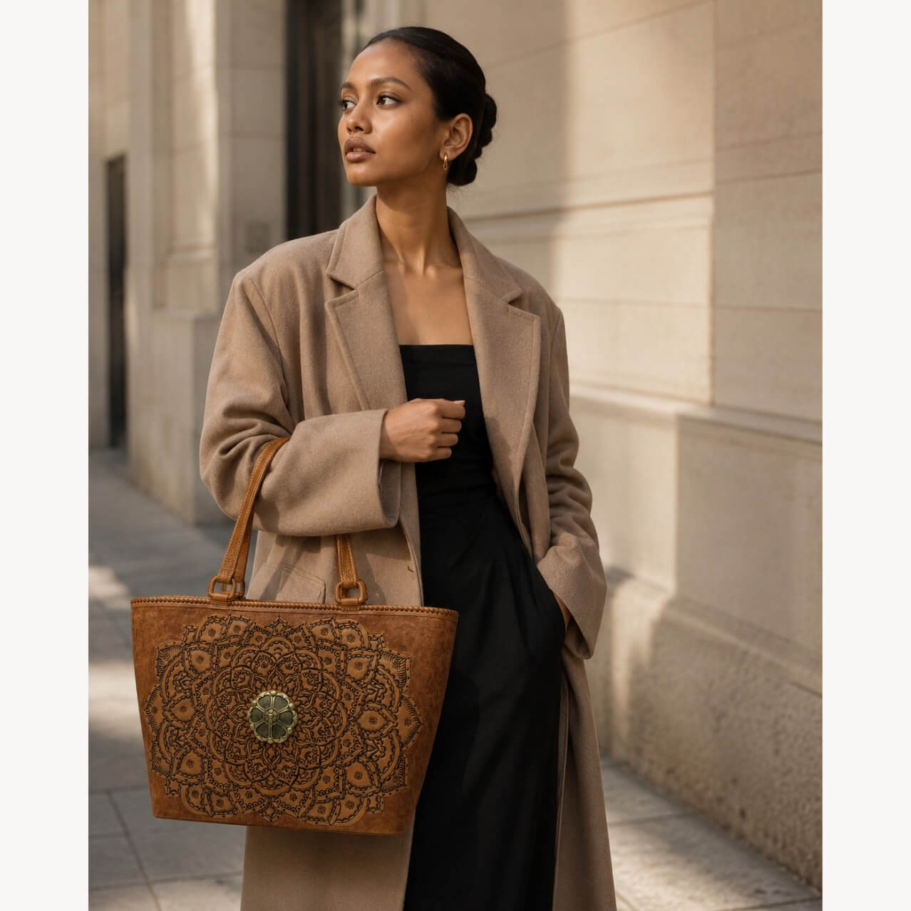 Cognac/burnished brown nude color tooled leather tote with floral medallion hardware carried by a model in a camel coat on a city sidewalk.