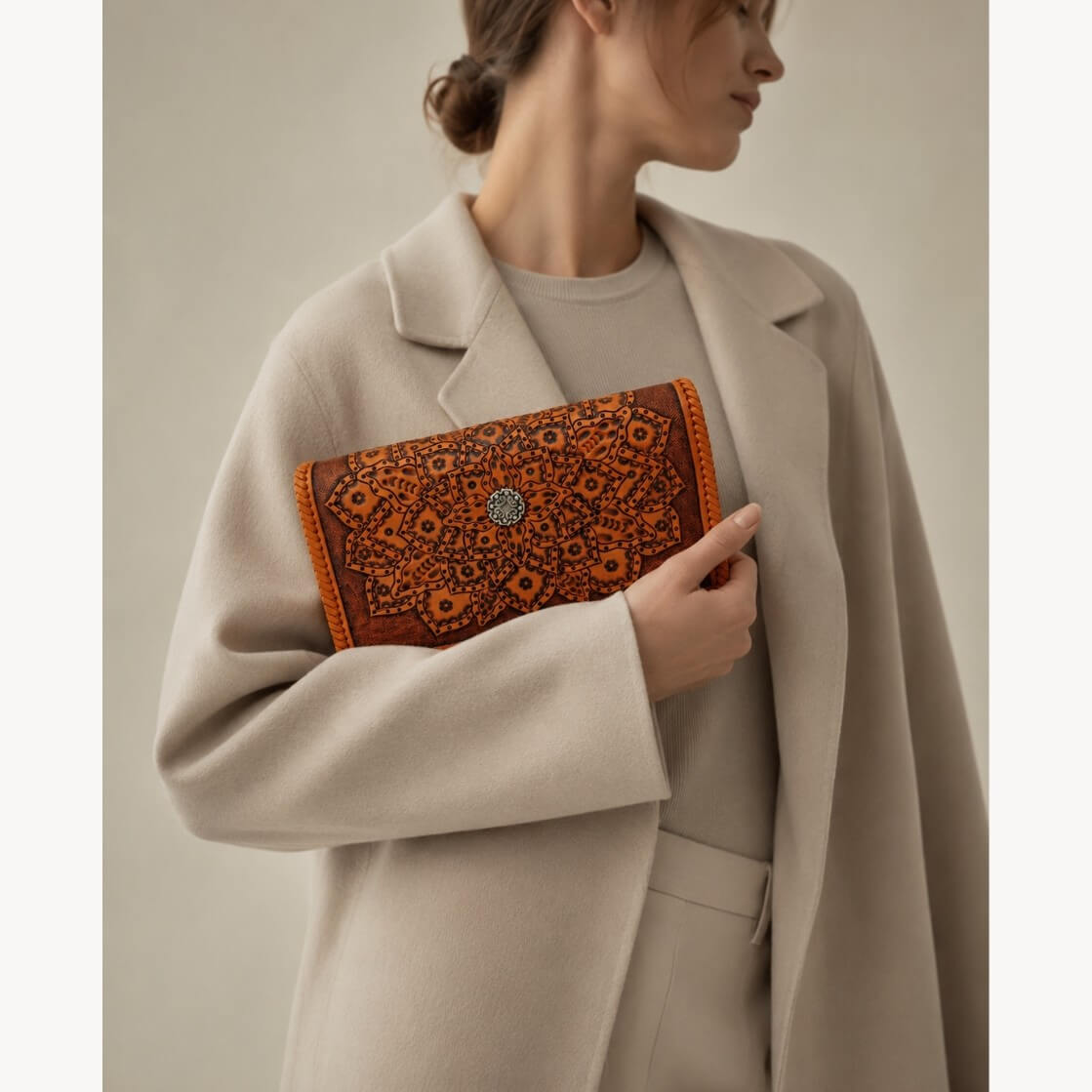 Model in a beige coat holding a cognac embossed leather clutch with mandala detailing, braided edges, and a circular metal clasp.