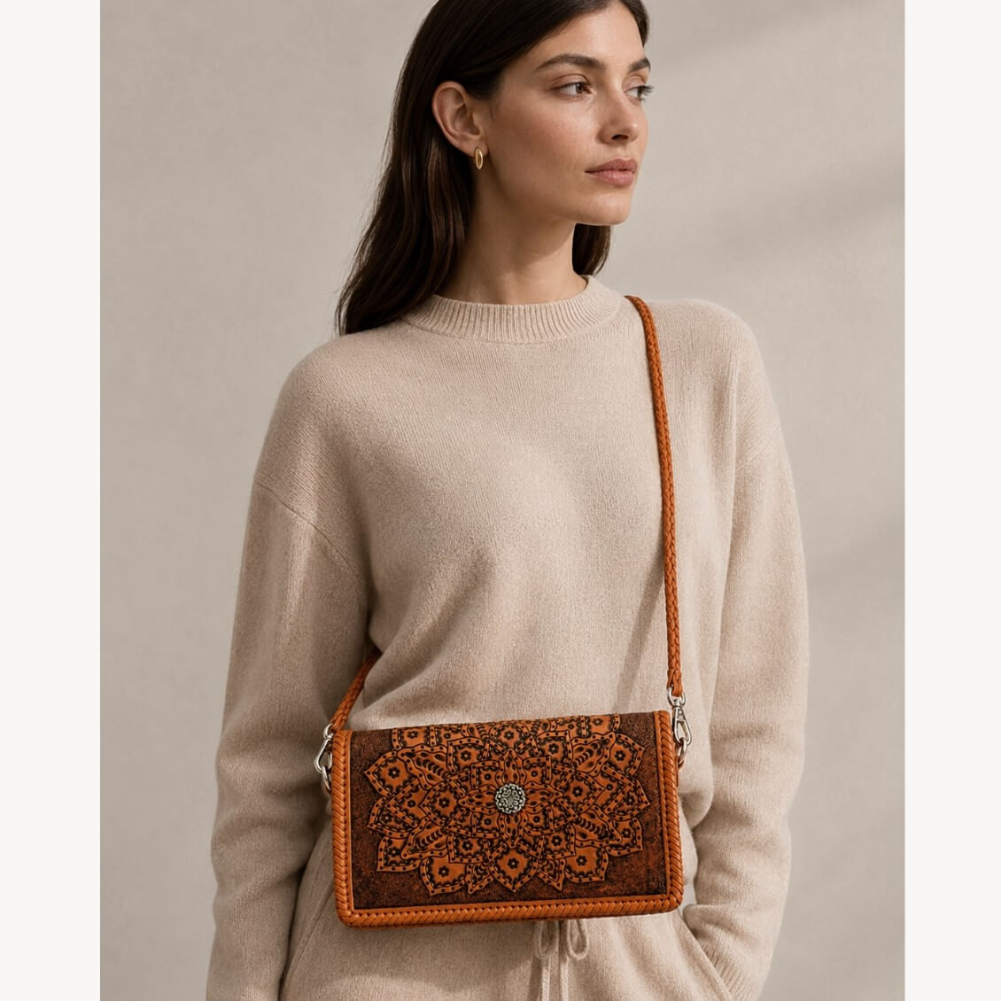 Model in a beige sweater wearing a cognac embossed leather crossbody bag with mandala detailing, braided strap, silver hardware, and a circular clasp.