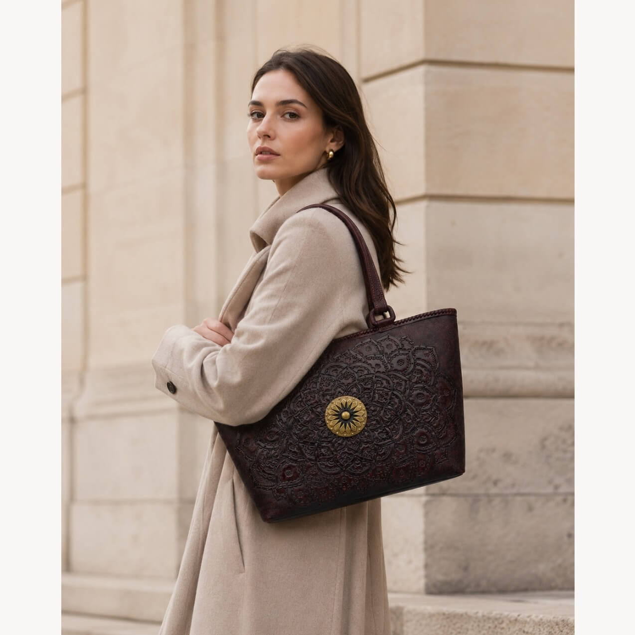 Model wearing a beige coat carrying a mahogany espresso embossed leather tote with top handles and a round gold medallion.