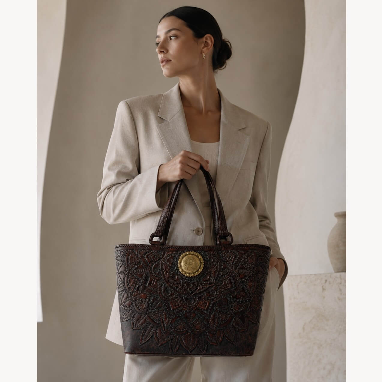 Model wearing a beige suit carrying a mahogany espresso floral embossed leather tote with top handles and a round gold medallion.