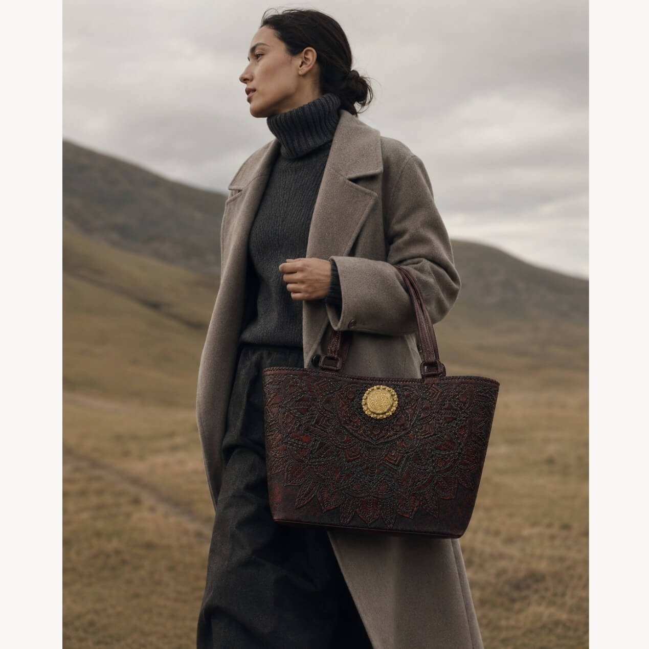Model wearing a gray coat carrying a mahogany espresso floral embossed leather tote with top handles and a round gold medallion in an outdoor landscape.