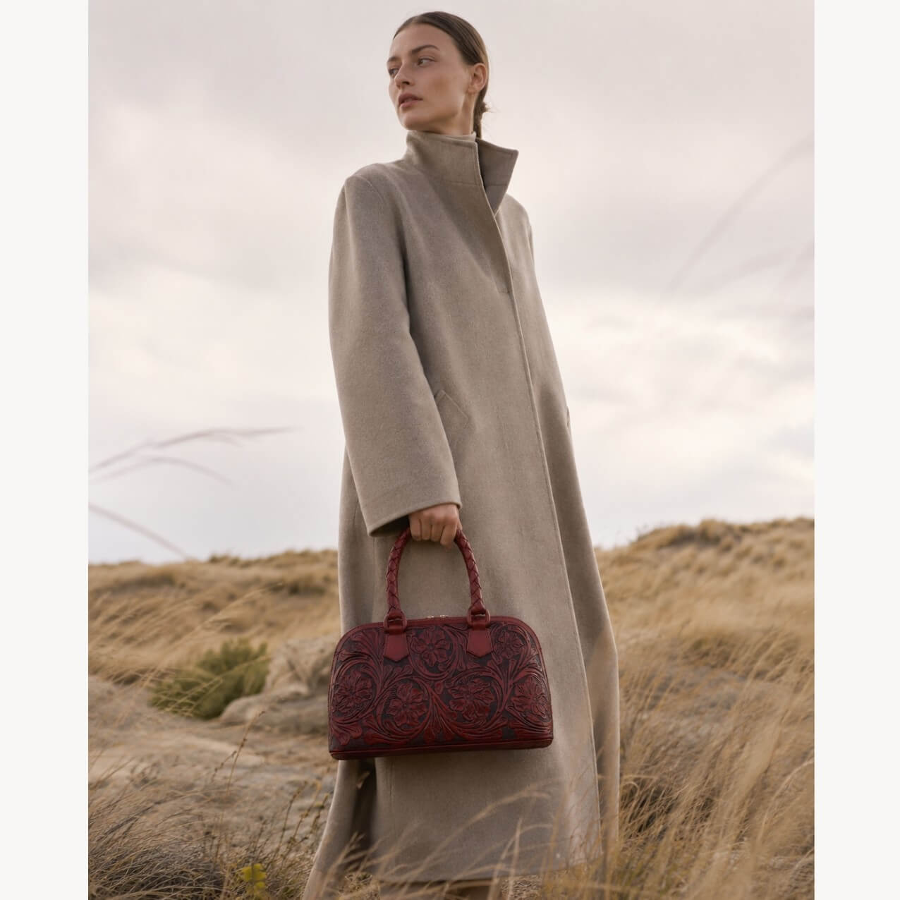 Model holding a Mahogany Espresso hand-tooled leather satchel bag with floral detailing and braided top handles.