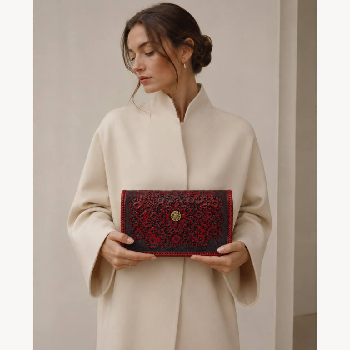 Model in a cream coat holding a Mahogany Espresso embossed leather clutch with mandala detailing, braided edges, and a gold circular clasp.