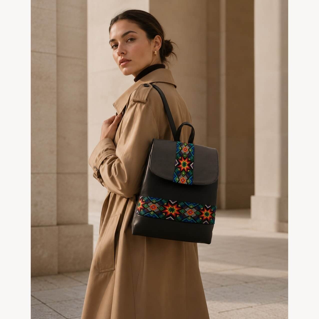 Model wearing a black leather backpack with colorful woven geometric textile panels over a tan trench coat in a modern architectural setting.