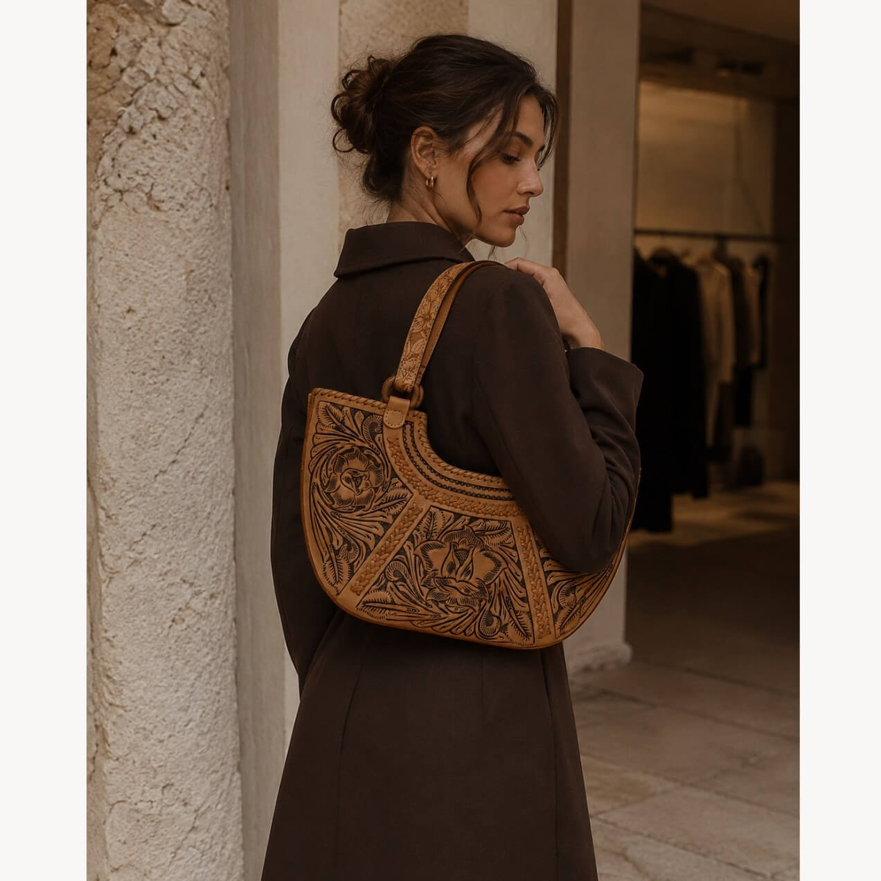 Model wearing a natural tan hand-tooled floral leather shoulder bag with carved floral detailing, styled with a brown coat in a stone architectural setting.