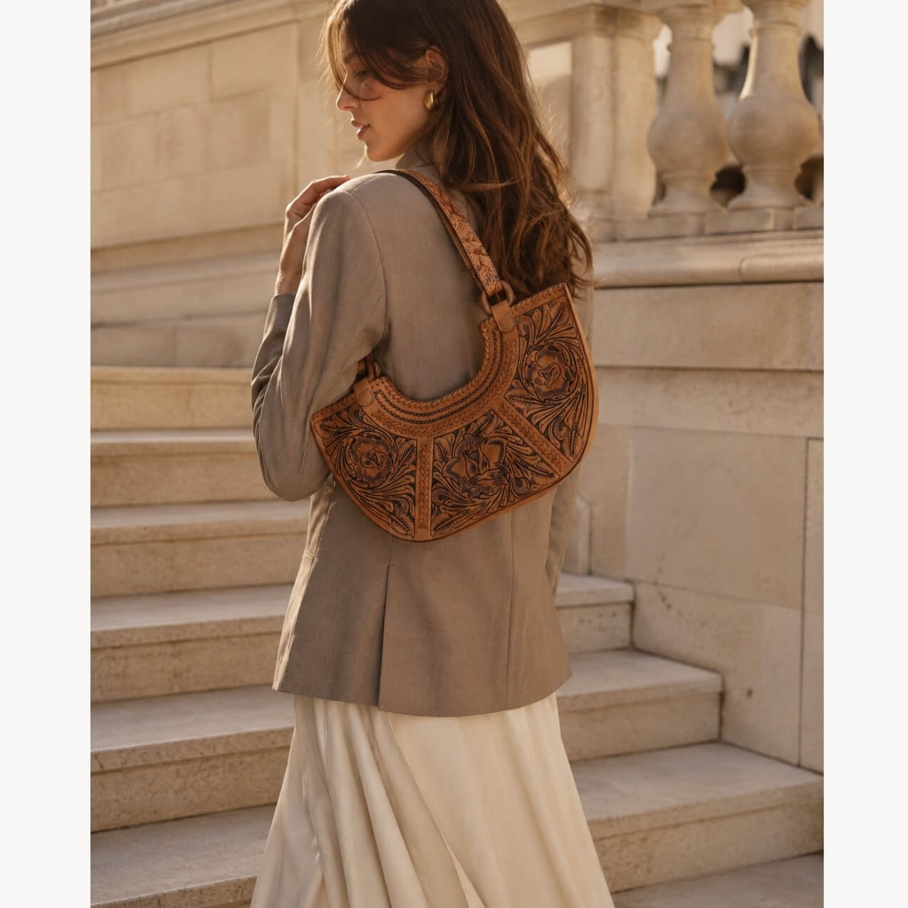 Model wearing a natural tan hand-tooled floral leather shoulder bag with carved floral detailing, styled with a gray blazer and cream skirt on stone steps.