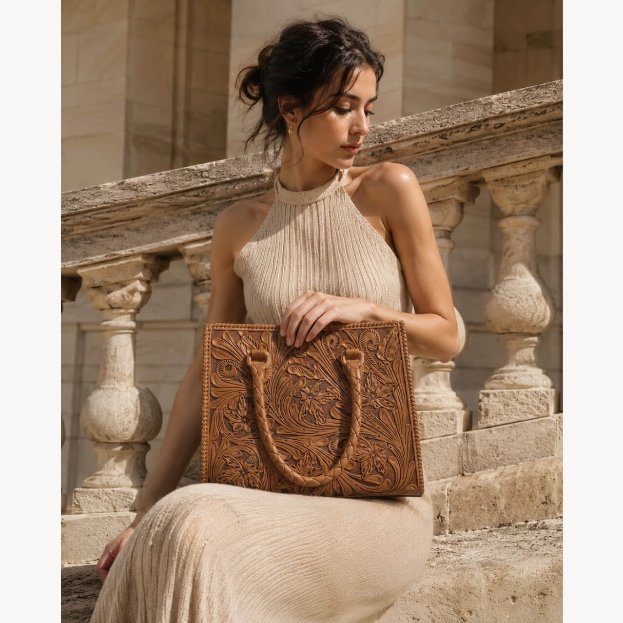 Model holding a natural tan hand-tooled floral leather tote bag with braided handles, styled with a cream knit dress in a stone architectural setting.
