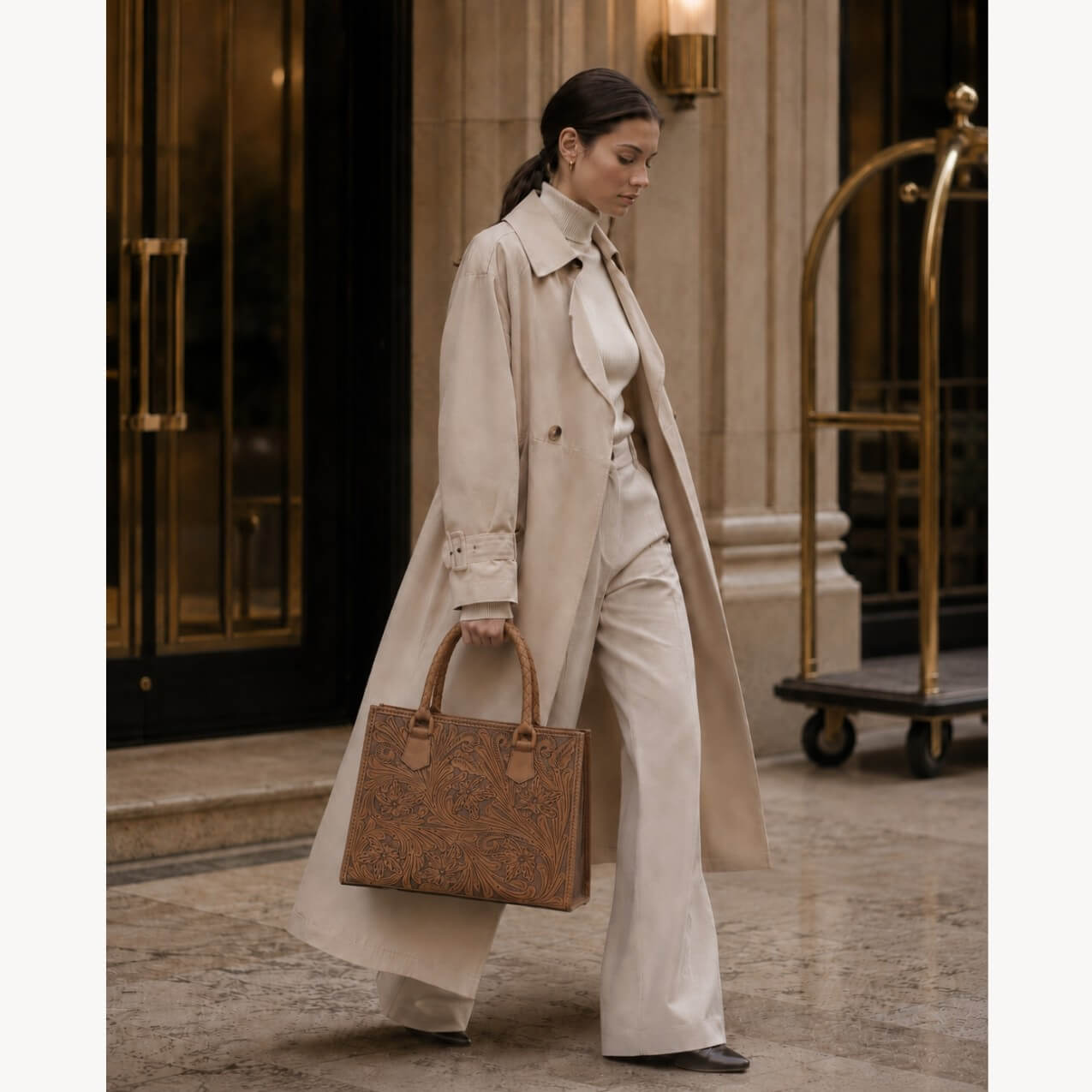 Model carrying a natural tan hand-tooled floral leather tote bag with braided handles, styled with a cream trench coat in a luxury architectural setting.