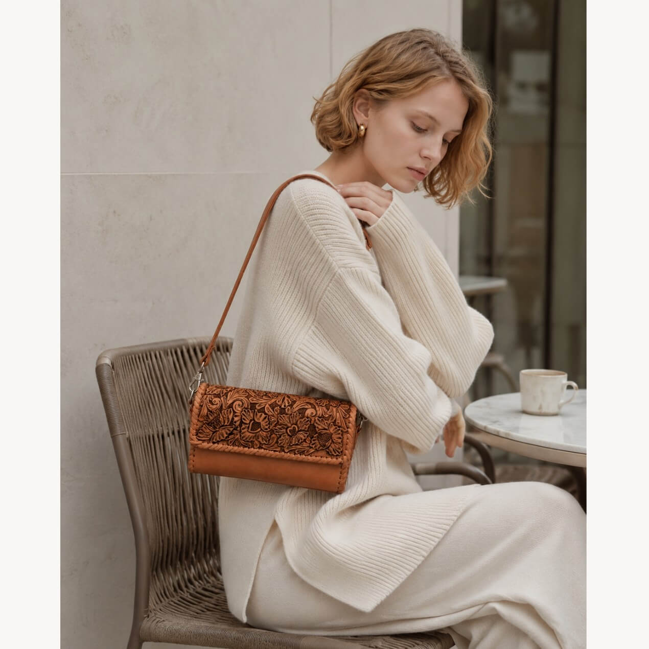 Seated model wearing a natural tan hand-tooled leather crossbody bag with floral detailing and a slim leather shoulder strap.