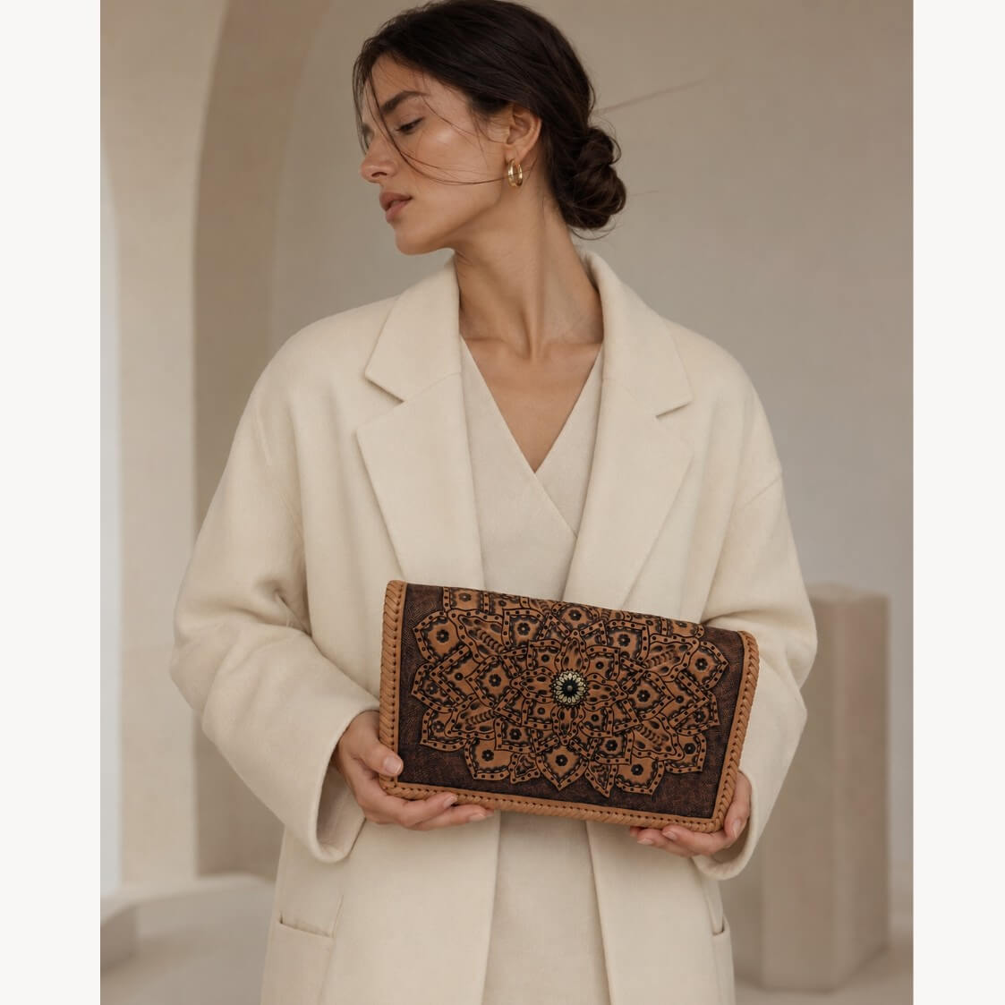 Model in a cream coat holding a natural tan embossed leather clutch with mandala detailing, braided edges, and a circular clasp.
