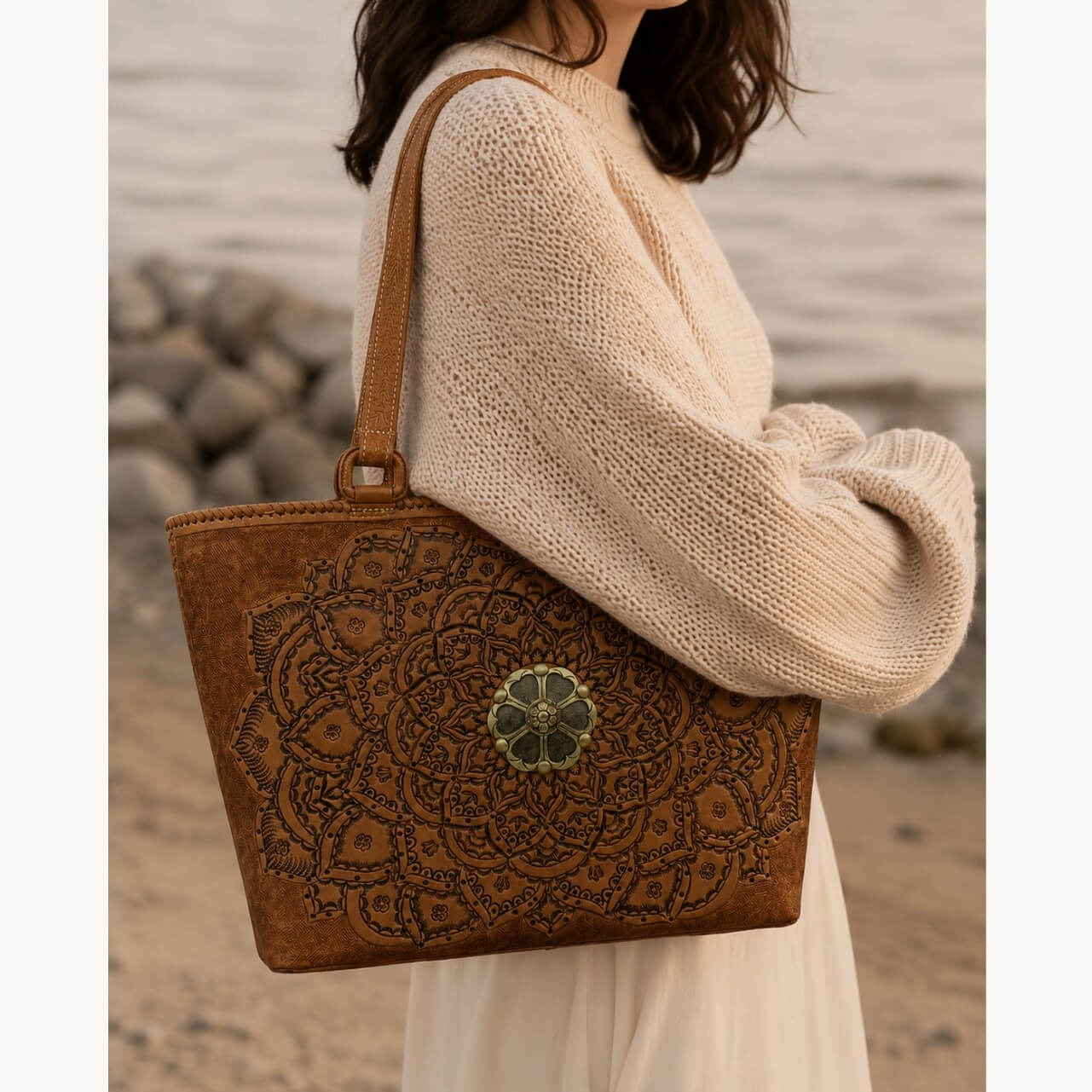 Cognac/burnished brown nude color tooled leather tote shoulder bag with floral medallion detail carried by a model in a cream sweater near the beach.