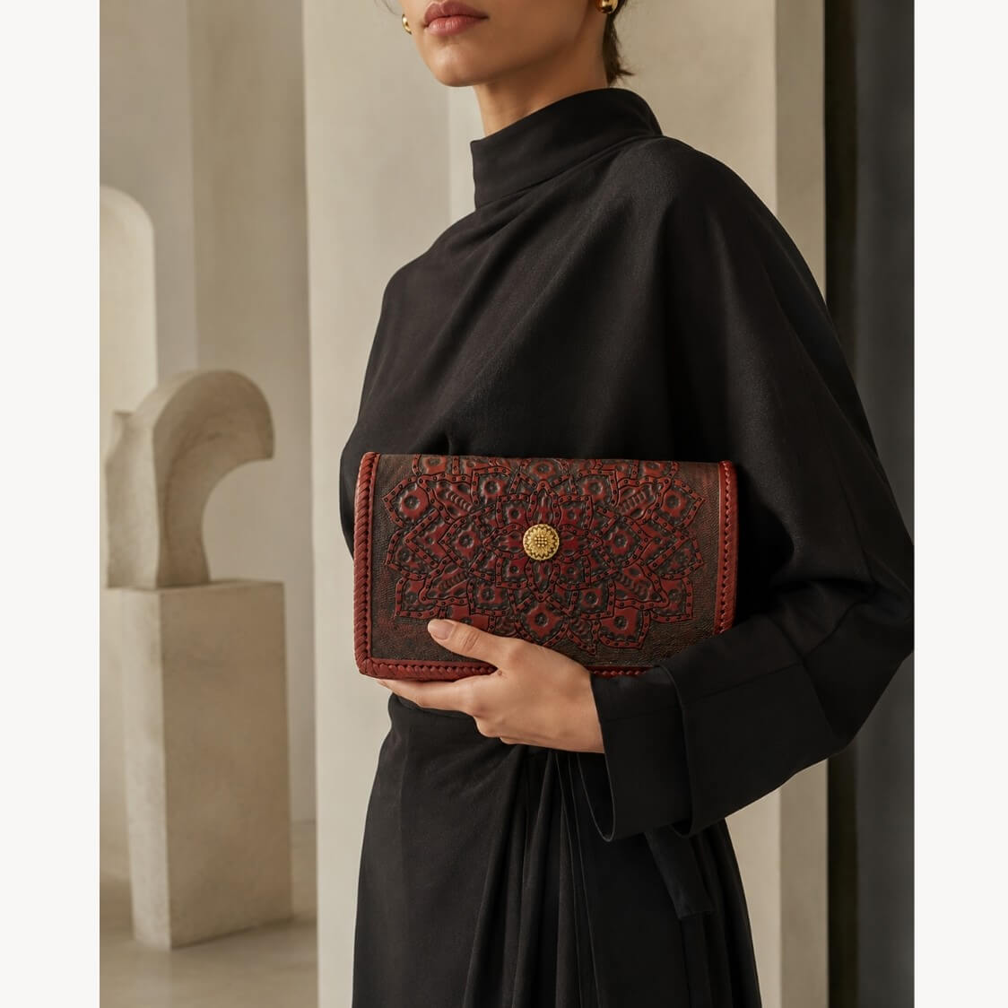 Model in a black dress holding a red-brown embossed leather clutch with mandala detailing and a gold circular clasp.
